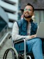 Man in wheelchair with laptop on lap in modern office outdoor setting, smiling.