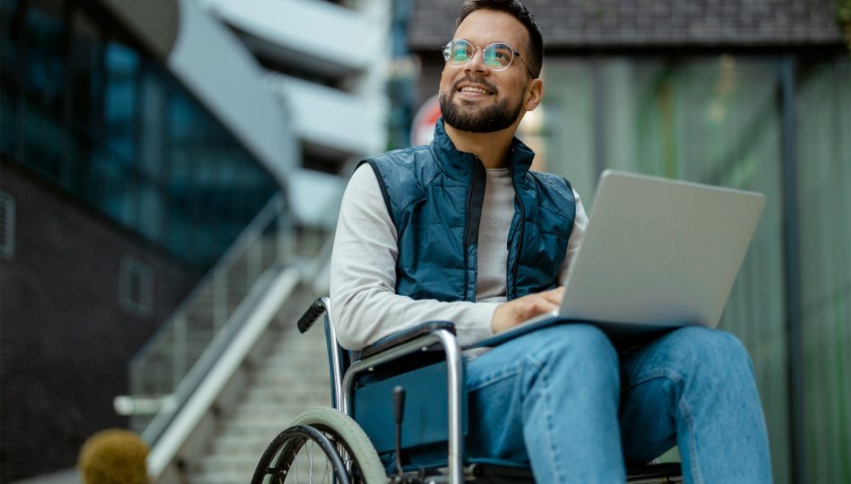 Man in wheelchair with laptop on lap in modern office outdoor setting, smiling.